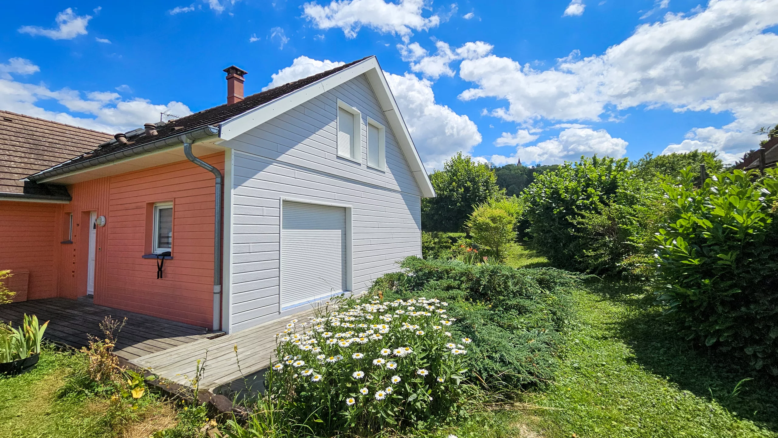Façade d'une maison moderne bicolore avec bardage en bois saumon et gris, une terrasse, et un jardin verdoyant sous un ciel bleu ensoleillé.